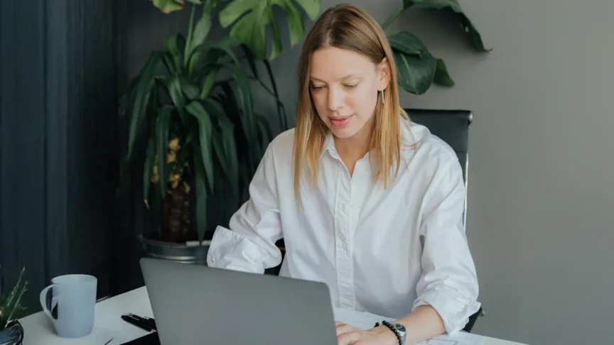 Woman in a white shirt working on a laptop at her desk with plants in the background.