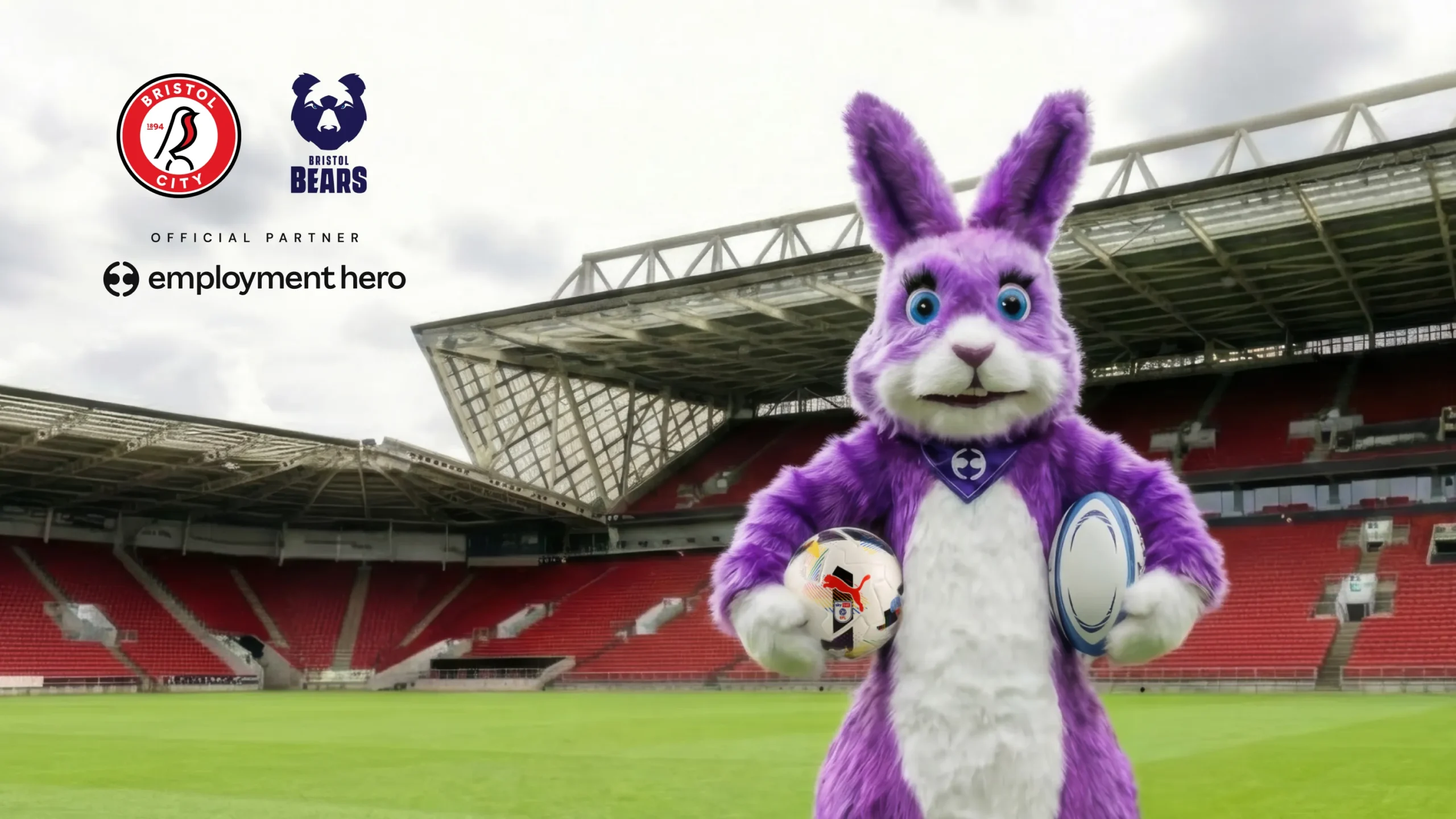 A purple rabbit mascot holding a soccer ball and a rugby ball in an empty sports stadium.